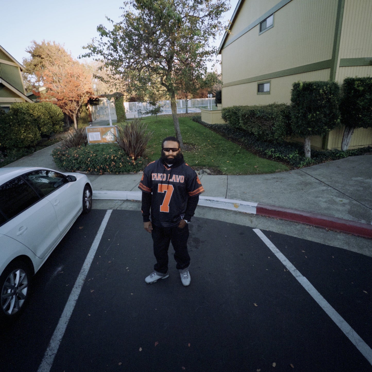 Person wearing a sports jersey standing in a parking lot with a building and trees in the background.