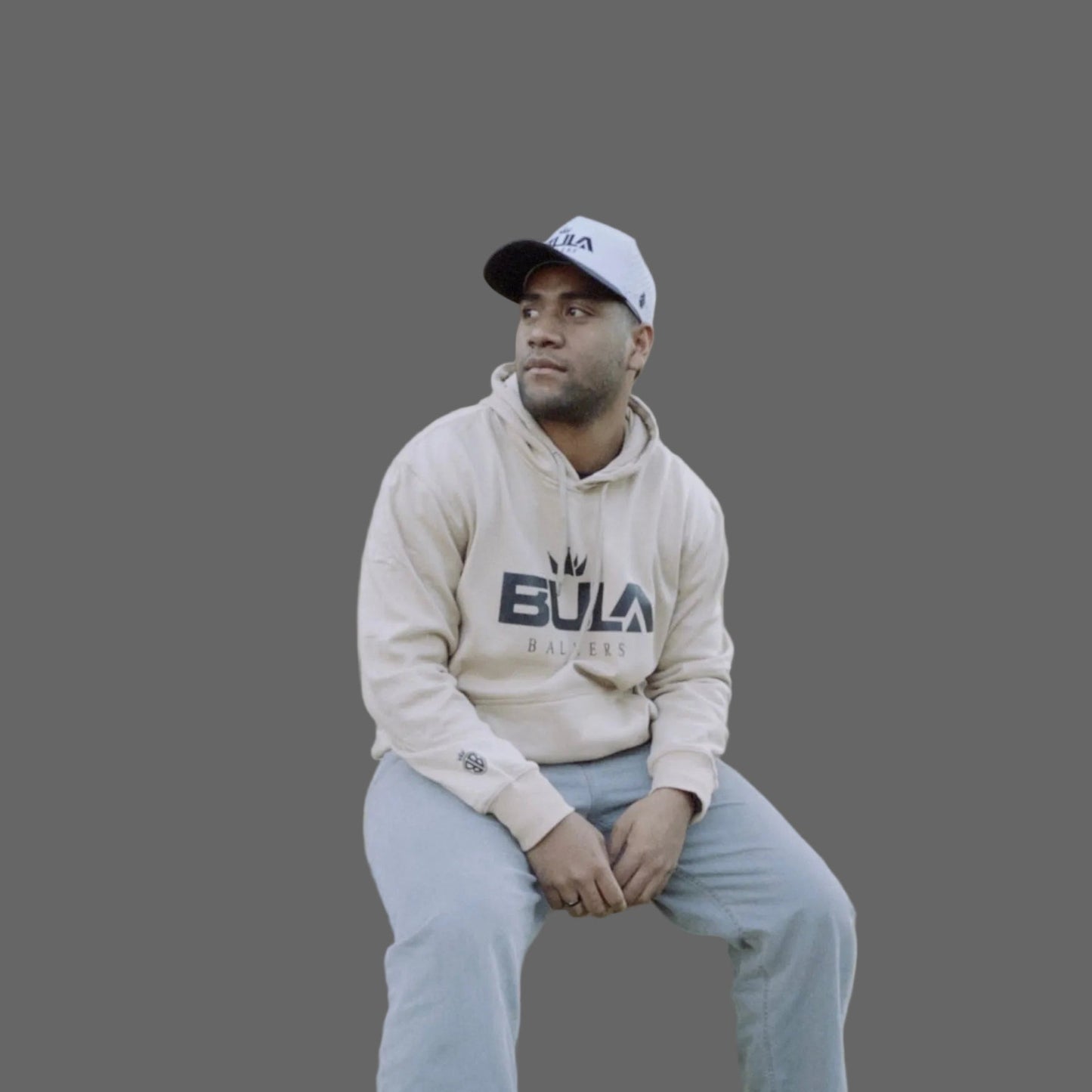 Person wearing a beige hoodie with 'Bula' branding and a cap, sitting on a gray background